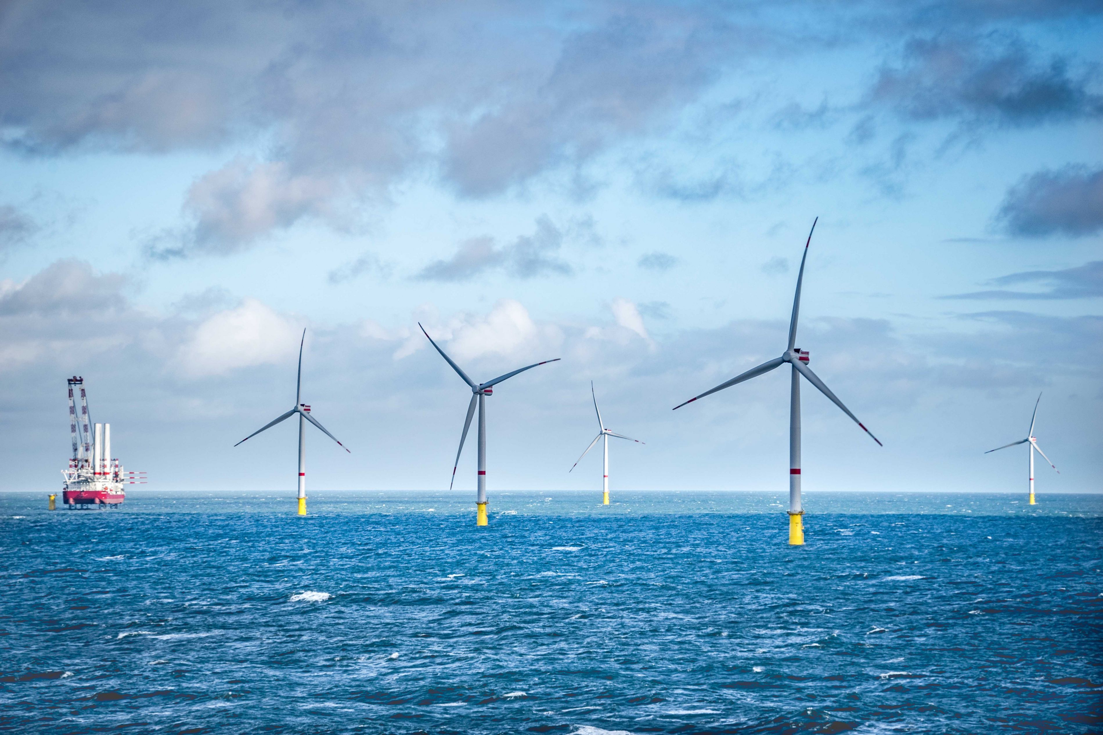 Five wind turbines in a body of water with a boat in the background - Becoming a rock solid investment for offshore wind energy.” investment for offshore wind energy